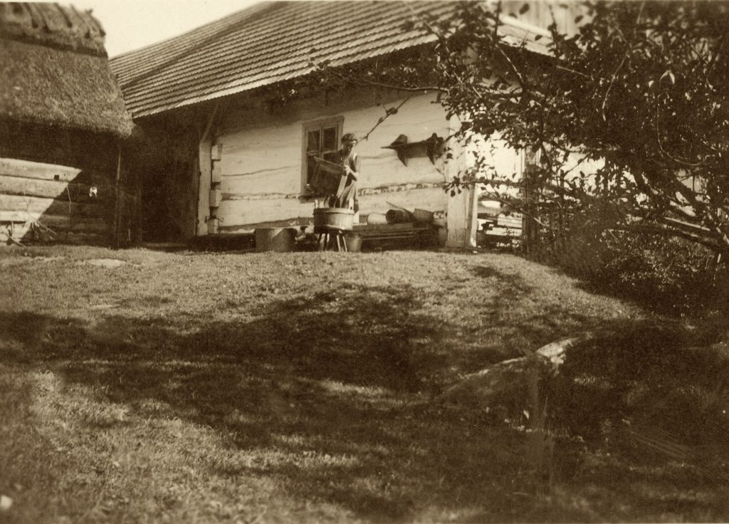 Archiwalne zdjęcie Chałupy Szylarów. Na pierwszym planie kobieta oporządzająca ule. W tle dom. Po lewej stronie szopa. 