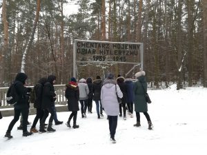 Grupa wchodzi na cmentarz przez bramę nad którą jest napis: Cmentarz Wojenny Ofiar Hitleryzmu 1939-1945. Zimowa aura.