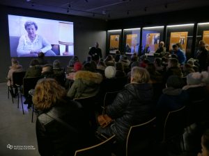 Grupa osób siedzi w sali czasowej Muzeum Polaków Ratujących Żydów. Na ścianie wyświetlany jest film. Z prawej strony kilka gablot ekspozycyjnych ściennych z planszami wystawy.