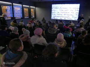 Grupa osób siedzi w sali czasowej Muzeum Polaków Ratujących Żydów. Na ścianie wyświetlany jest film. Z lewej strony kilka gablot ekspozycyjnych ściennych z planszami wystawy.