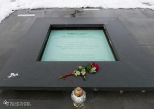 Zdjęcie współczesne. Sceneria zimowa. Na pierwszym planie pomnik w kształcie płyty poświęcony pamięci żydowskich ofiar Zagłady i ich anonimowych polskich wspomożycieli, na którym umieszczono znicz i różę.