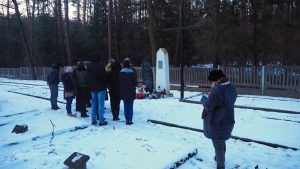 Grupa stoi przed pomnikiem na cmentarzu. Na pierwszym planie fragmenty nagrobków. W tle drzewa. Zimowa aura.
