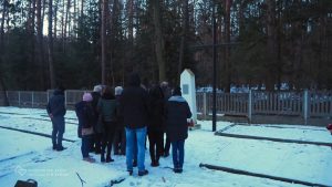 Grupa stoi przed pomnikiem na cmentarzu. Na pierwszym planie fragmenty nagrobków. W tle drzewa. Zimowa aura.