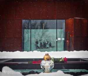 Zdjęcie współczesne. Sceneria zimowa. Na pierwszym planie pomnik w kształcie płyty poświęcony pamięci żydowskich ofiar Zagłady i ich anonimowych polskich wspomożycieli, na którym umieszczono znicz i różę. W tle fasada Muzeum.
