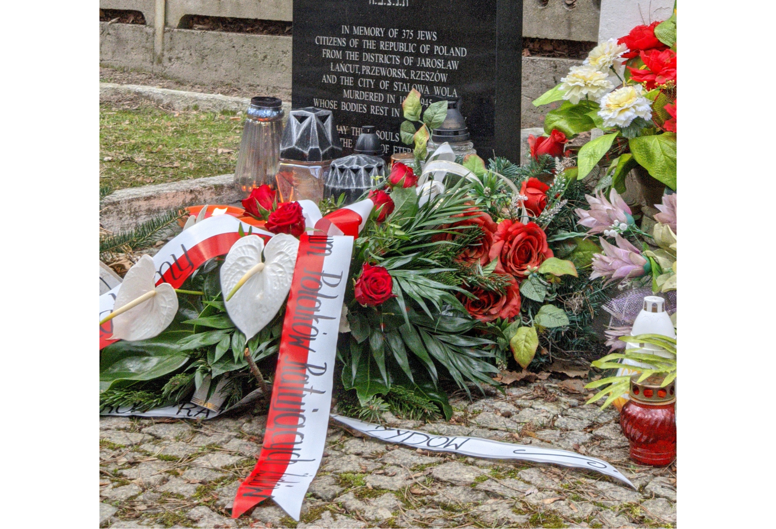 Zdjęcie. Zbliżenie na wiązankę kwiatów złożoną przed pomnikiem. Kwiaty i wstęgi w barwach biało-czerwonych.