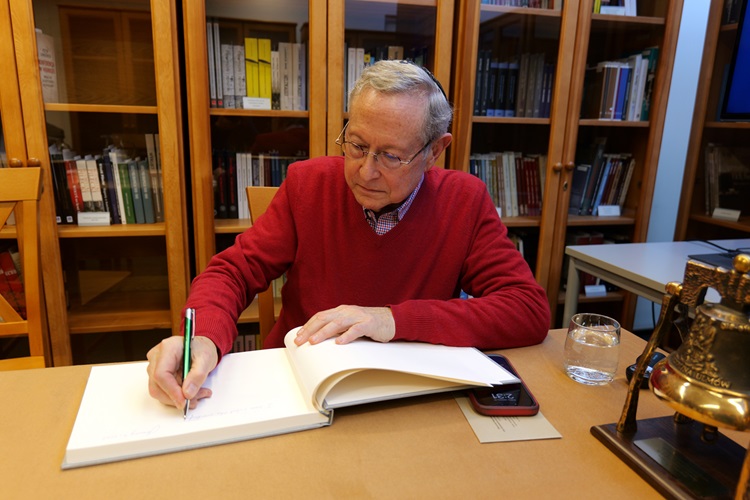 Jeffrey Cymbler siedzi przy biurku i dokonuje wpisu do pamiątkowej księgi. W tle biblioteka.