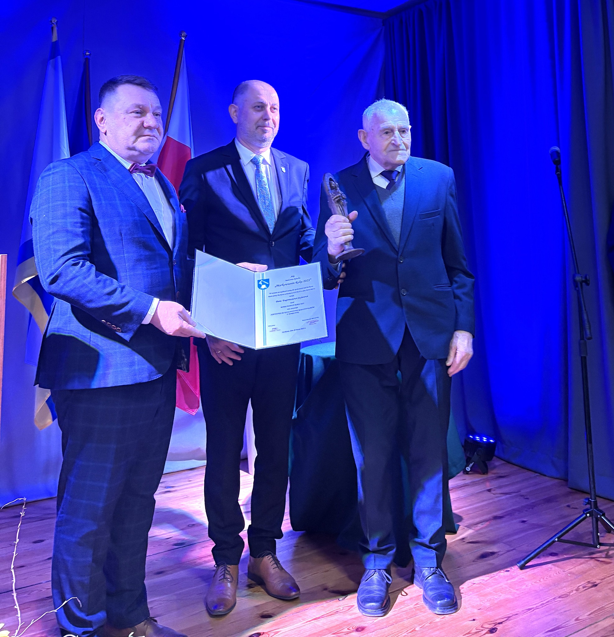 Na zdjęciu widzimy laureata, który stoi na scenie w towarzystwie dwóch mężczyzn. Jeden z nich trzyma w ręku dyplom, a drugi statuetkę.