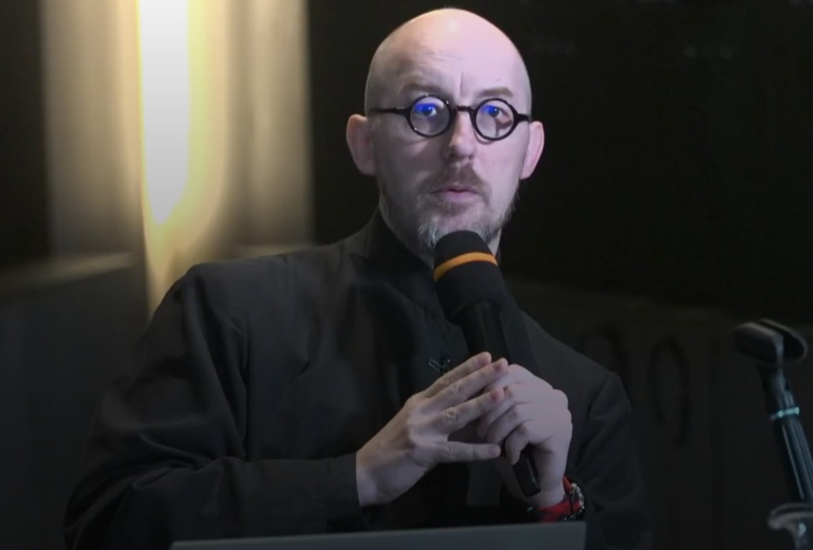 Prelegent siedzi przy biurku, na tle ekspozycji muzealnej.