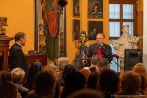 Waldemar Rataj przemawia do publiczności w Muzeum Archidiecezjalnym w Przemyślu. W tle obrazy.
