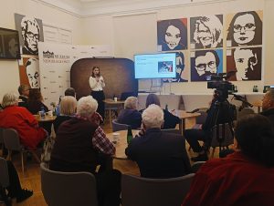 Prelegentka stoi przed biurkiem. Po prawej stronie rzutnik z prezentacją.