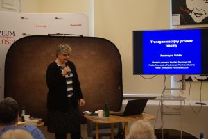 Prelegentka stoi przed biurkiem. Po prawej stronie rzutnik z prezentacją.