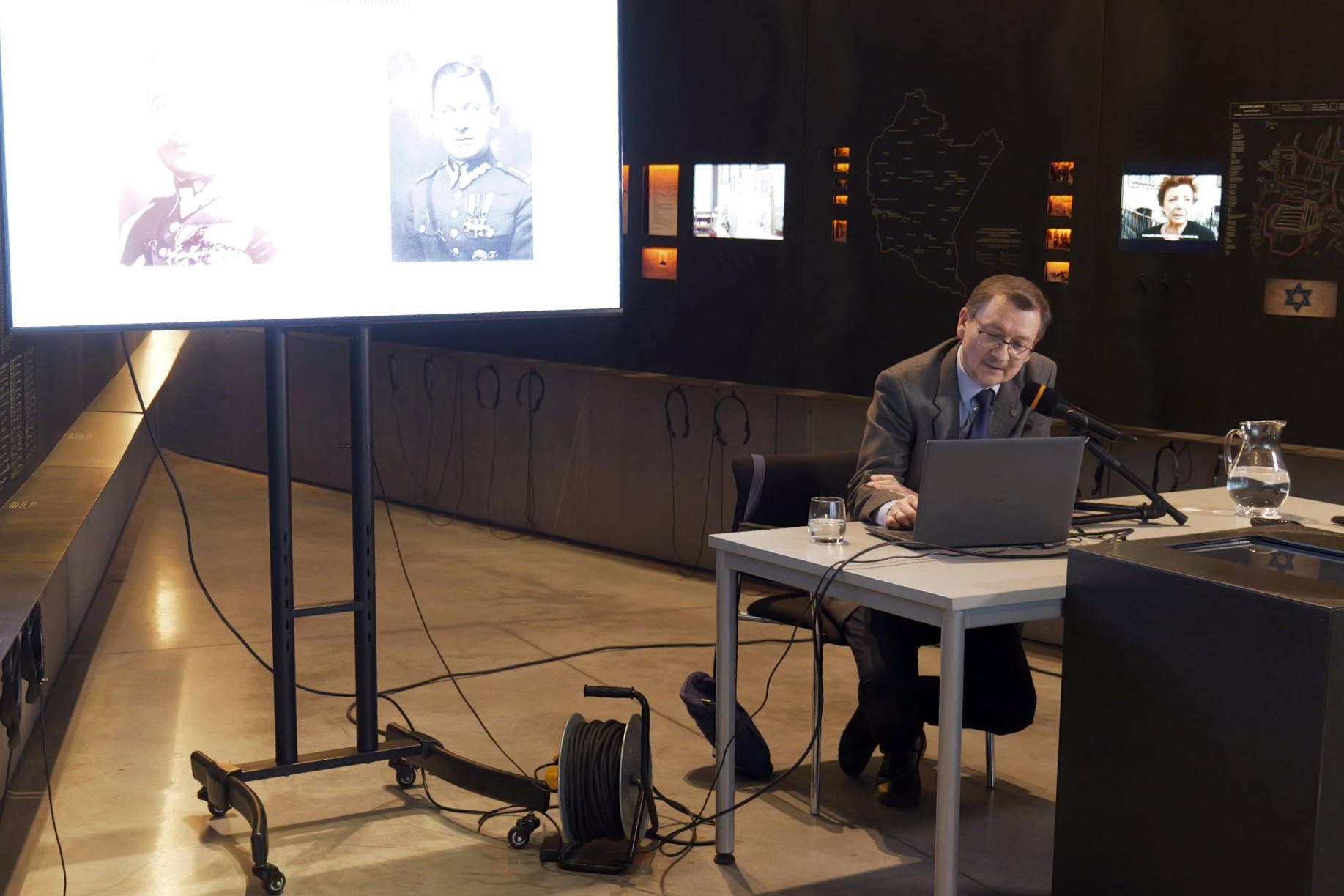 Prelegent siedzi przy biurku. W tle ekspozycja muzealna oraz z lewej strony rzutnik.