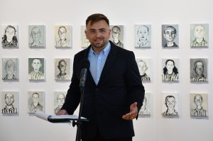 Prelegent przemawia do publiczności. W tle obrazy olejne.