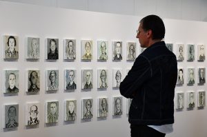 Mężczyzna zwiedzający wystawę, przyglądający się obrazom więźniów KL Prelegent przemawia. W tle obrazy więźniów KL Auschwitz.