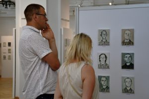 Mężczyzna i kobieta zwiedzający wystawę, przyglądający się obrazom więźniów KL Prelegent przemawia. W tle obrazy więźniów KL Auschwitz.