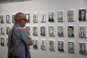 Mężczyzna zwiedzający wystawę, przyglądający się obrazom więźniów KL Prelegent przemawia. W tle obrazy więźniów KL Auschwitz.