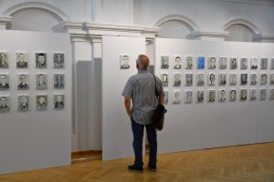 Mężczyzna zwiedzający wystawę, przyglądający się obrazom więźniów KL Prelegent przemawia. W tle obrazy więźniów KL Auschwitz.