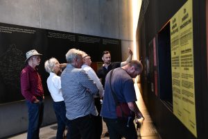 Grupa osób zwiedza ekspozycję muzealną z przewodnikiem.