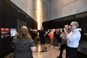 Grupa osób zwiedza ekspozycję muzealną z przewodnikiem.