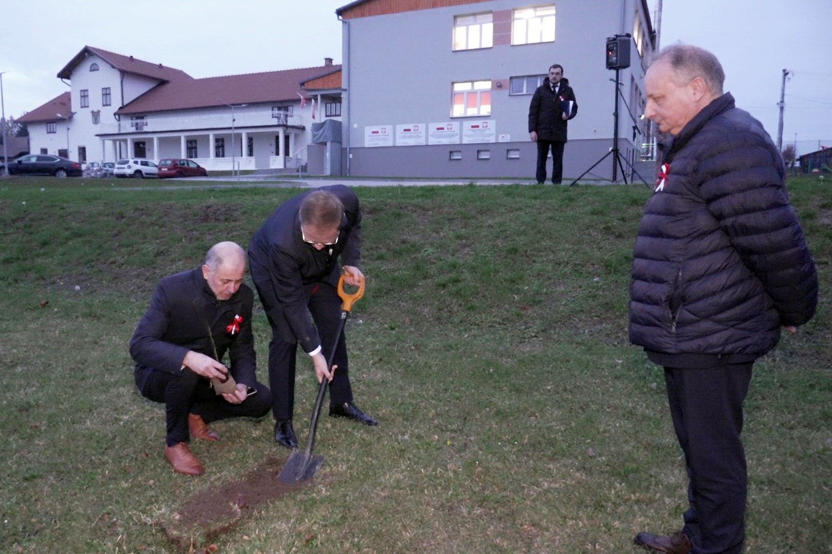 Trzech mężczyzn sadzących drzewko. W tle budynek.